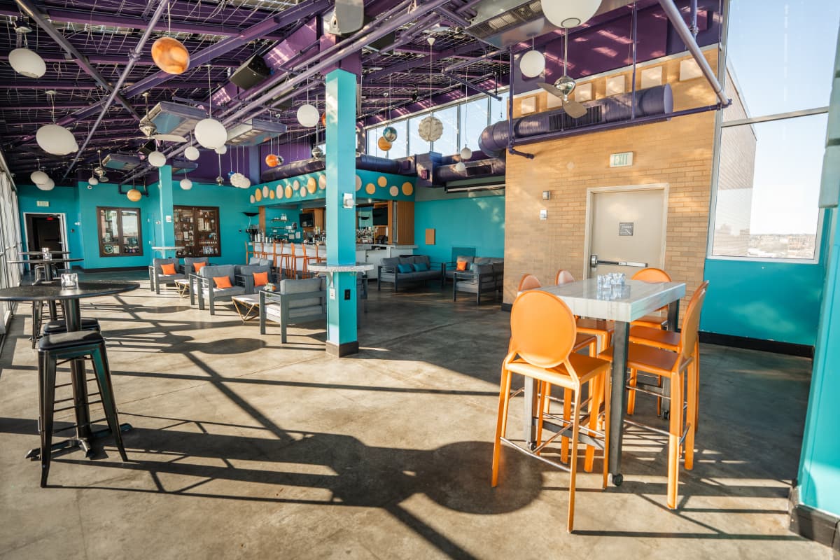 A view of all of the seating options in the twilight room. Tall bar tables with orange seats with back support. Tall black bar tables with backless barstools. Several couches dispersed throughout. Tall seats line the bar.