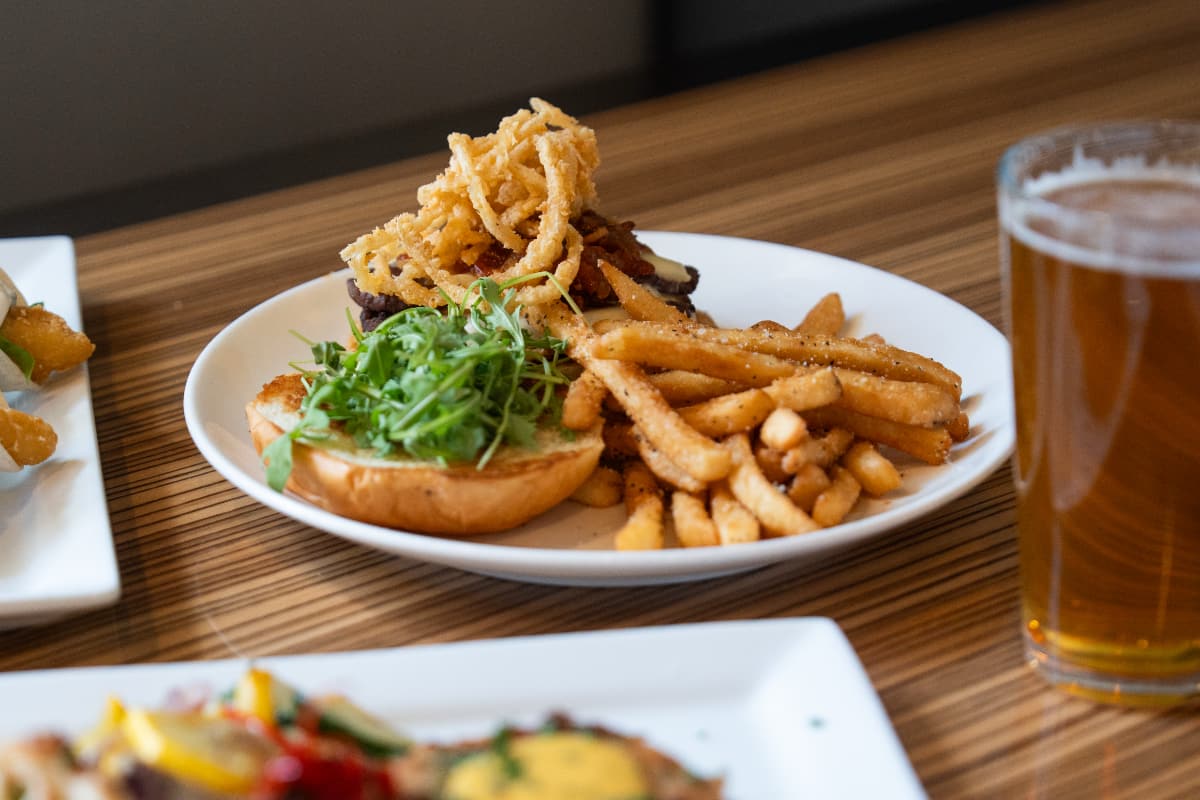 A focus on a burger topped with greens and flash fried onions. Fries are on the plate, and a beer is next to the plate. Other plates of food are blurred in the foreground.