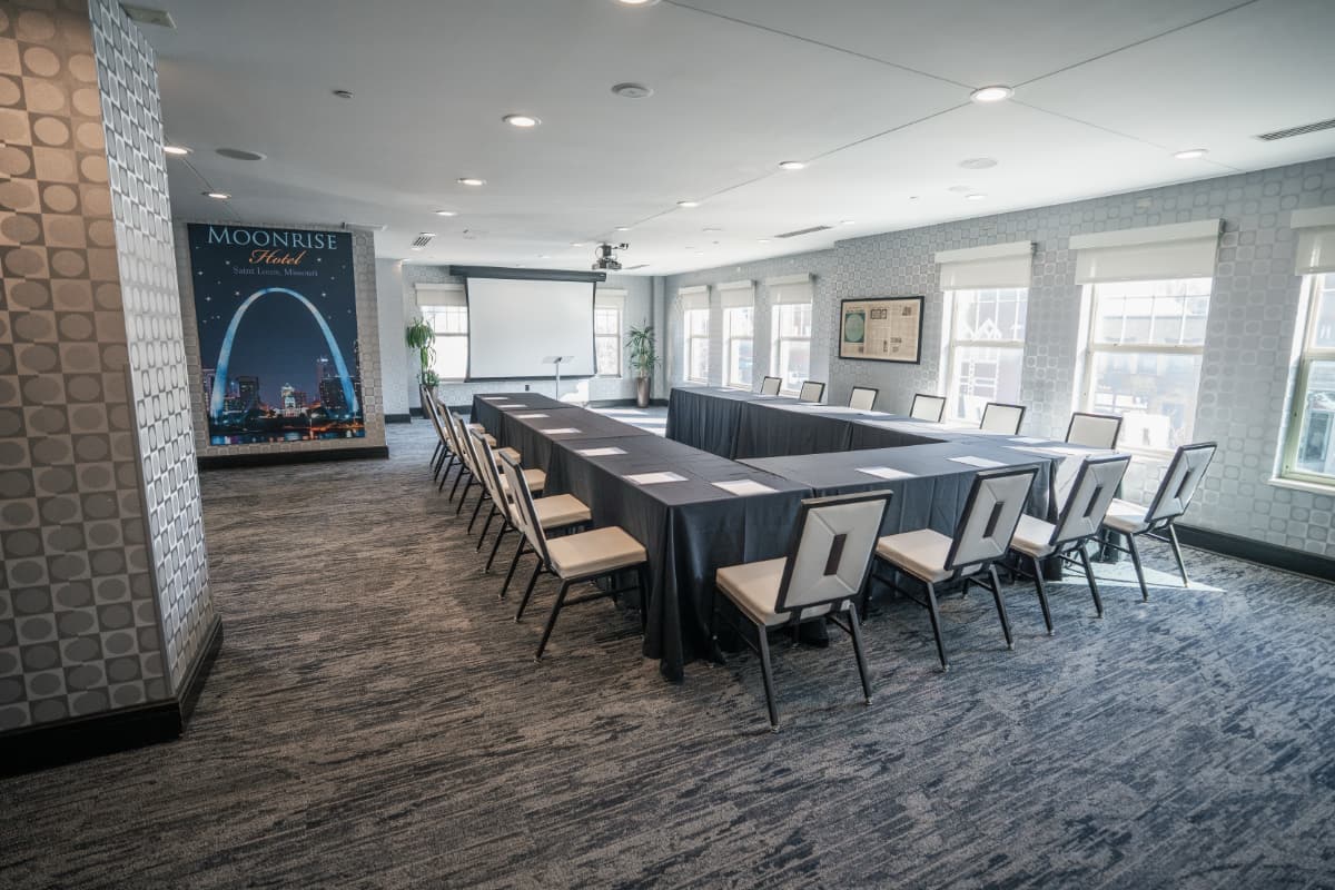 meeting room Meeting tables shaped in a U-formation surrounded by chairs. A projector screen hangs from the ceiling. A giant art piece of the Arch hang on the wall off to the side.