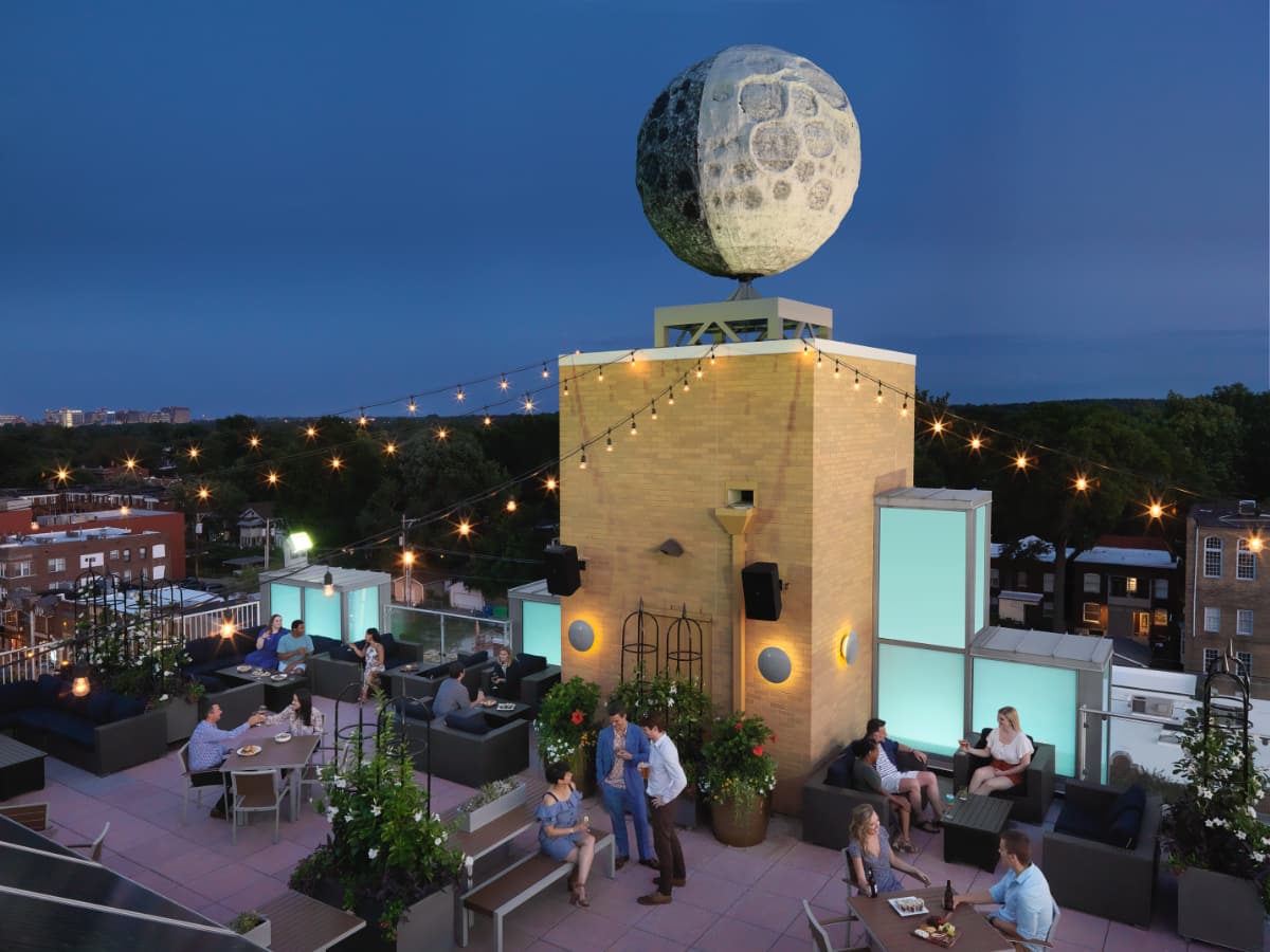 rooftop garden bar at dusk A high up shot of the Rooftop Garden Bar at dusk. People socialize beneath the illuminated moon statue and decorative lights glow.